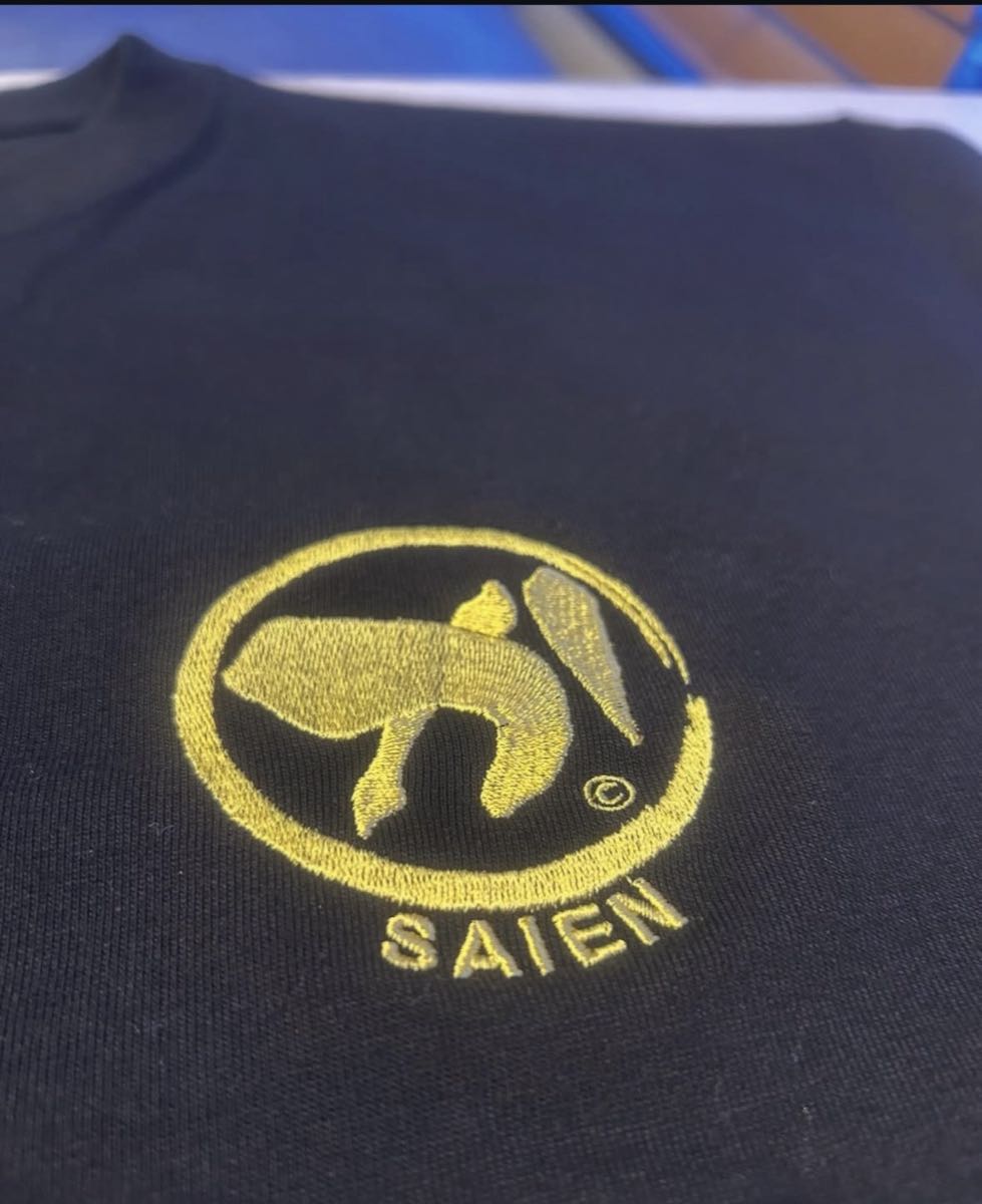 黒いTシャツの胸元に入れた金糸の丸型ロゴ刺繍のクローズアップ